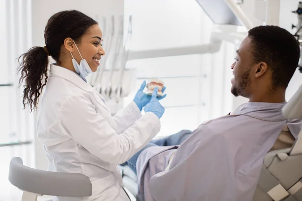 Χαμογελώντας Black Stomatologist Lady Consulting Αρσενικό ασθενή στην κλινική, κρατώντας πλαστικό σαγόνι — Φωτογραφία Αρχείου