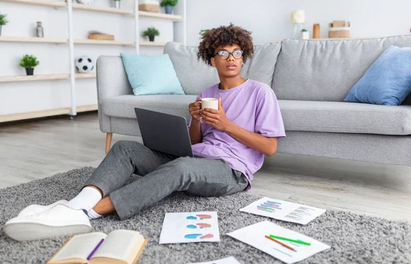 Adolescente preto pensativo sentado no chão com laptop e materiais de estudo, fazendo pausa, bebendo café em casa — Fotografia de Stock