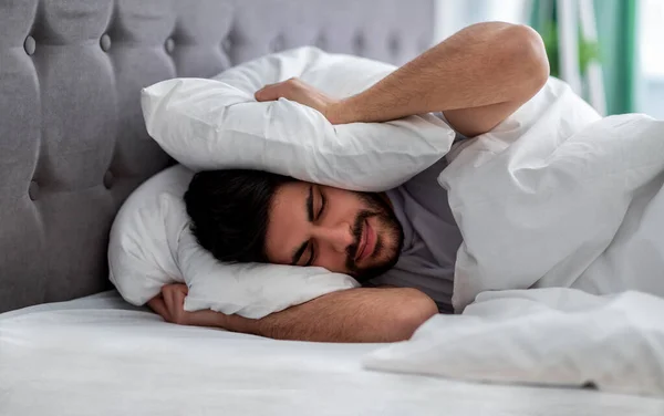 Hombre árabe trastornado cerrando sus oídos con almohada, escondiéndose del sonido o ruido demasiado fuerte temprano en la mañana, acostado en la cama —  Fotos de Stock