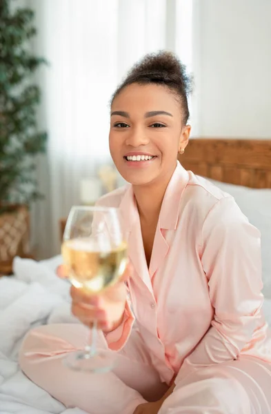 Time for youself concept. Portrait of happy african american lady sitting on bed, drinking wine and smiling at camera — Stock Photo, Image