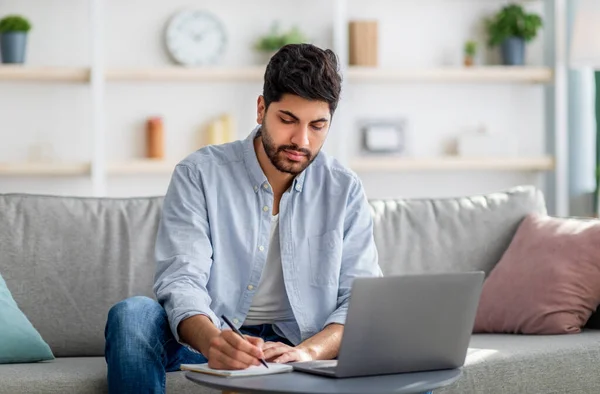 아랍 남성 프리랜서는 노트를 작성하고 노트북을 사용하며 소파에 앉아 있는 동안 노트를 쓰고 컴퓨터를 사용 한다 — 스톡 사진