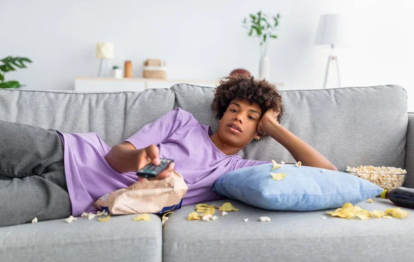 Lui zwart tiener guy liggend op bank met verspreid voedsel houden tv-controller, kijken saaie film op televisie thuis — Stockfoto