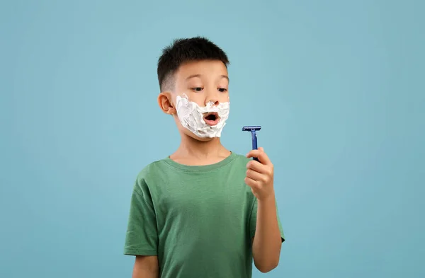 Schockiert kleine asiatische Junge mit Rasierschaum im Gesicht Blick auf Rasiermesser — Stockfoto