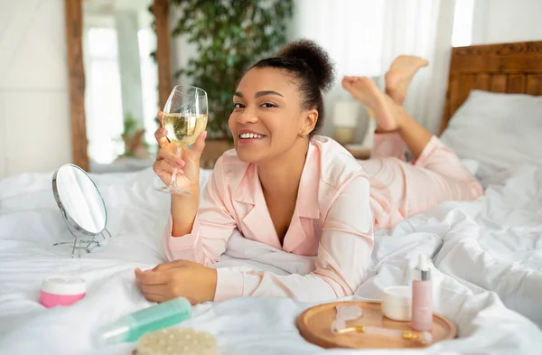 Jong gelukkig afrikaanse amerikaanse dame in slaapkledij genieten wijn liggend op bed met beauty care producten naast haar — Stockfoto