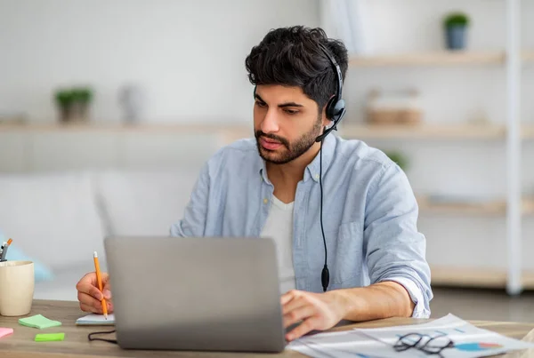 Heureux homme d'affaires arabe dans les écouteurs ayant une réunion d'affaires en ligne sur ordinateur portable, écrire des informations, assis au bureau à la maison — Photo