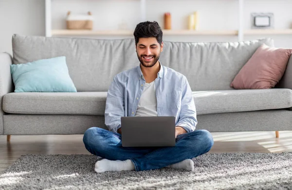 Arab millennial człowiek za pomocą laptopa, przeglądanie Internetu, pracy online podczas siedzenia na podłodze w domu — Zdjęcie stockowe