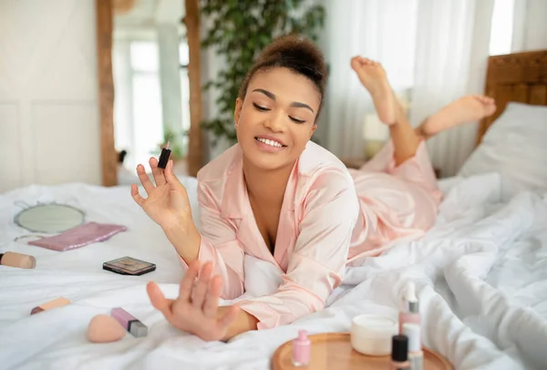 Ganska afrikansk amerikansk kvinna applicera nagellack, göra manikyr när du ligger på sängen hemma — Stockfoto