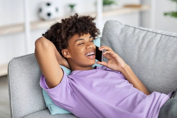 Веселый черный подросток лежит на диване, разговаривает по телефону с другом дома — стоковое фото