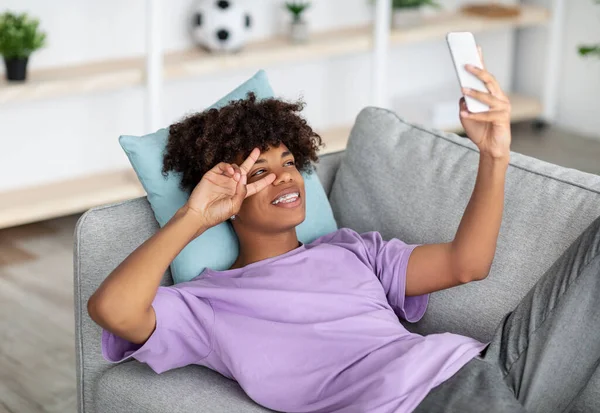 Positiv svart tonåring kille som ligger på soffan, tar selfie eller gör videosamtal på smartphone hemifrån — Stockfoto