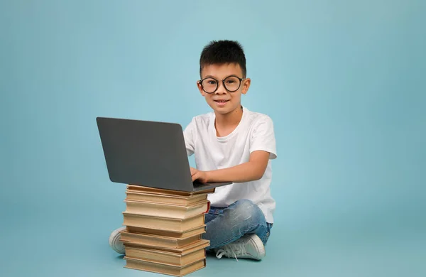 Mały azjatycki chłopiec w okularach siedzi z laptopem i stos książek — Zdjęcie stockowe