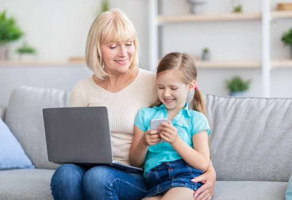 Happy mature woman using laptop hugging girl with smartphone — Stock Photo, Image