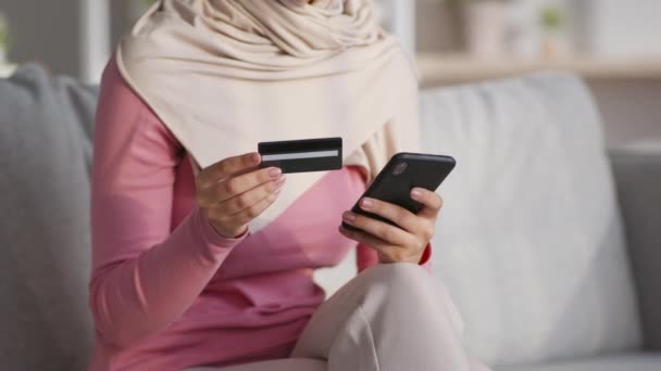 Zakupy online. Nierozpoznawalna muzułmanka dokonująca transakcji internetowej w domu, korzystająca z karty kredytowej i telefonu komórkowego — Wideo stockowe