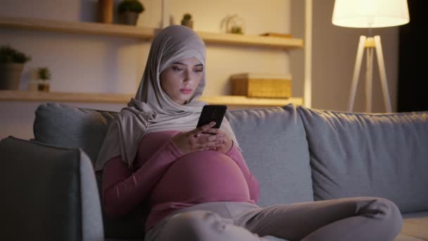 어린 임산부가 소파에 앉아서 스마트폰으로 뉴스나 아기보는 법을 인터넷에서 읽고 있습니다 — 비디오