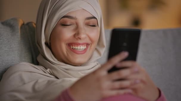 Televisión digital en el bolsillo. Joven mujer musulmana alegre en hijab viendo una película en un teléfono inteligente y riendo por la noche en casa — Vídeo de stock