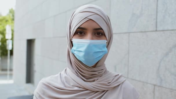 Mujer musulmana joven en hijab fijando máscara médica protectora en su cara, mirando a la cámara al aire libre, seguimiento de disparo — Vídeos de Stock