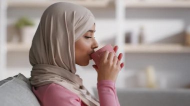 Ev eğlencesi konsepti. Huzurlu bir Arap kadının portresi. Sıcak çay içiyor, evdeki kanepede dinleniyor.