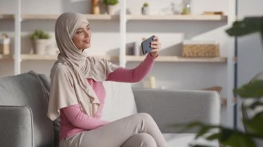 Başörtülü genç, pozitif ortadoğulu bayan cep telefonuyla selfie çekiyor ve evde sosyal medya için içerik oluşturuyor.