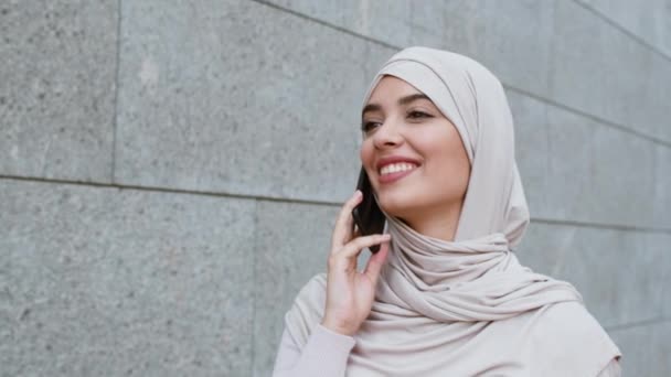 Хорошая мобильная связь. Молодая мусульманка, одетая в традиционный хиджаб, разговаривает по телефону и улыбается, гуляет на улице — стоковое видео