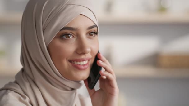 Mujer joven de mediana edad positiva que usa hijab hablando por teléfono celular con familiares o amigos, sentada en casa, espacio vacío — Vídeos de Stock