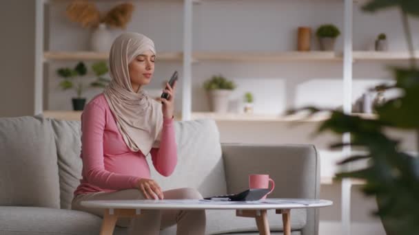 Graviditet og fritid. Muslimsk gravid dame i traditionelt tørklæde hviler på sofaen derhjemme og besvarer telefonopkald – Stock-video