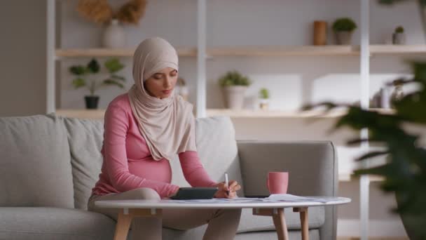 Young pregnant muslim woman writing check list at home, calculating expenses before childbirth, sitting on sofa at home — Stock Video