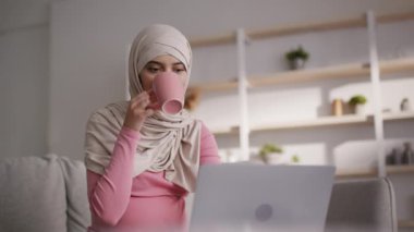 Sıcak bir iş yeri. Genç kaygısız orta doğulu kadın laptopla evde çalışıyor, sıcak çayın tadını çıkarıyor.