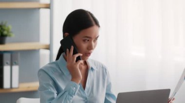 Çinli iş kadını cep telefonuyla konuşuyor laptopta oturuyor ve modern ofiste çalışıyor. Asyalı iş kadınının iş yerinde telefon görüşmesi yaparken yan görüntüsü. İletişim Kavramı