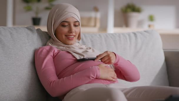 Έγκυος γυναίκα της Μέσης Ανατολής που αναπαύεται στον καναπέ και μιλάει σε smartphone με τηλέφωνο στην κοιλιά και χρησιμοποιώντας μεγάφωνο — Αρχείο Βίντεο