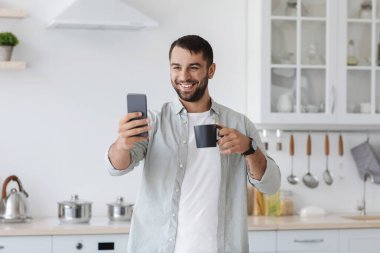 Sakallı neşeli yetişkin Avrupalı adam akıllı telefona bakıp kahve içiyor ve telefonda selfie çekiyor.