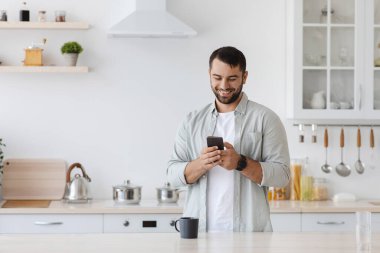 Gülümseyen beyaz adam, sakallı, akıllı telefondan mesaj yazıyor, sohbet ediyor ya da minimalist mutfak içinde blog yazıyor.