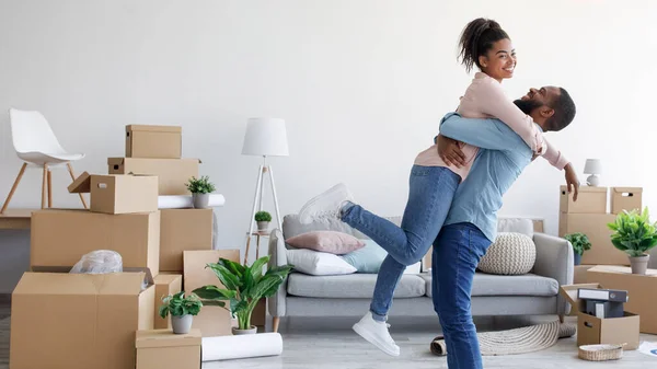 Feliz milenar afro-americano marido abraça e levanta sua esposa em novo apartamento em torno de caixas de papelão e coisas — Fotografia de Stock