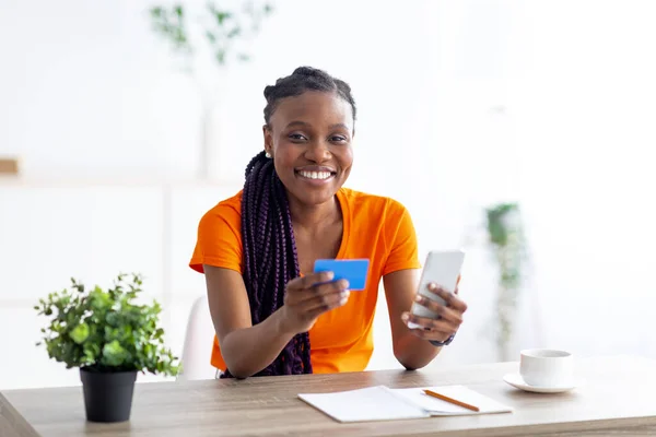Joven mujer afroamericana comprando en línea con tarjeta de crédito y teléfono móvil, sentado en el escritorio en el interior —  Fotos de Stock