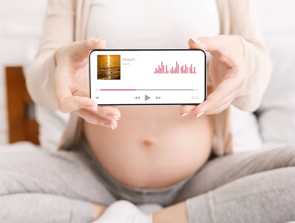 Pemain Musik Kehamilan. Wanita hamil memegang ponsel dengan antarmuka aplikasi pemutar audio — Stok Foto