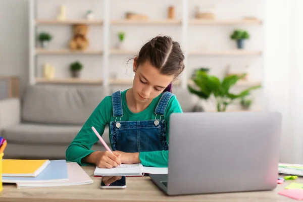 Chica sentada en el escritorio, usando el ordenador portátil, escribiendo en su libro de texto — Foto de Stock
