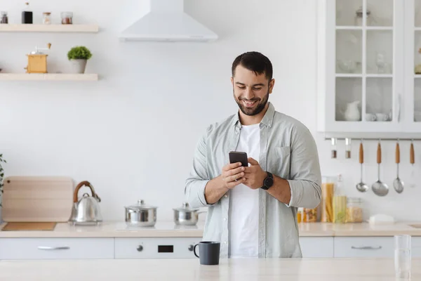 Sonriente hombre caucásico adulto con barba escribiendo mensaje en el teléfono inteligente, chat o blog en el interior de la cocina minimalista — Foto de Stock