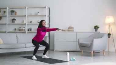 Sırt ve kalça egzersizi. Başörtüsü takan genç aktif Arap bayan evde derin çömelme egzersizi yapıyor, boş alan.