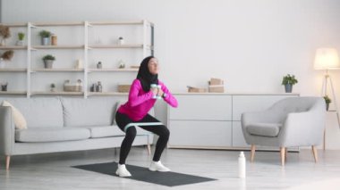 Genç aktif Müslüman kadın tesettür takıyor, evde elastik bant ve dambıl kullanarak derin çömelme egzersizleri yapıyor.