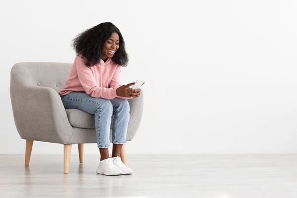 Mujer negra bonita positiva sentada en sillón con teléfono celular — Foto de Stock