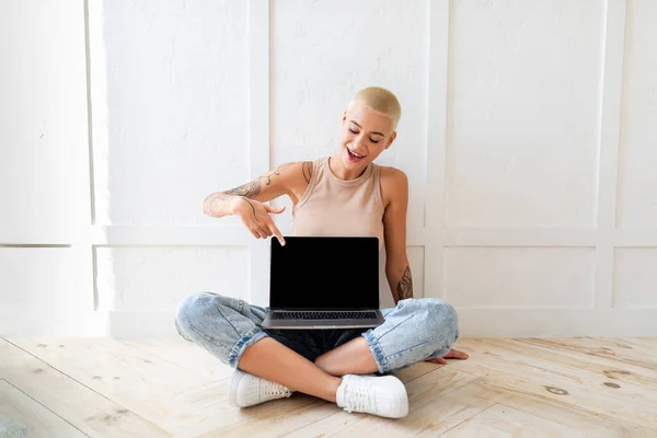 Wow, great website. Happy caucasian woman pointing at laptop with black screen, sitting on floor over light wall