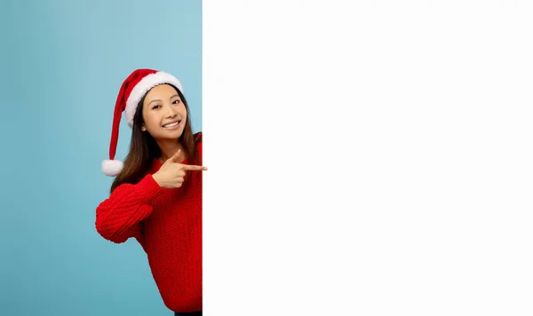 Vzrušená asijské dáma v santa klobouk ukazuje na prázdný banner s prostorem pro vaši reklamu přes modré pozadí, maketa pro design — Stock fotografie