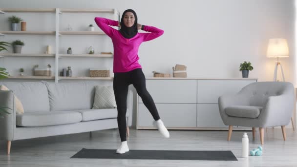 Щаслива спортивна середня східна леді в хіджабі працює вдома, практикуючи глибокі присідання і підняття ніг — стокове відео