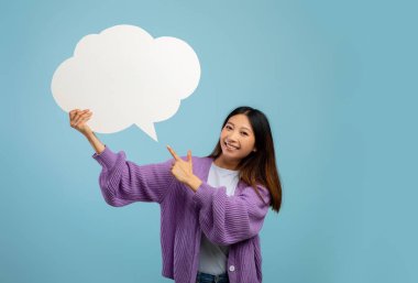 Boş konuşma balonunu işaret eden mutlu Asyalı kadın mavi stüdyo arka planındaki kameraya gülümsüyor.
