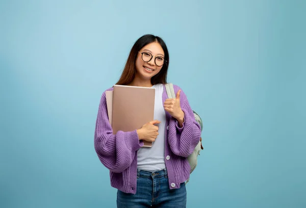 Pozytywna azjatycka studentka trzymająca zeszyty i pokazująca kciuk w górze, uśmiechnięta do kamery na niebieskim tle — Zdjęcie stockowe