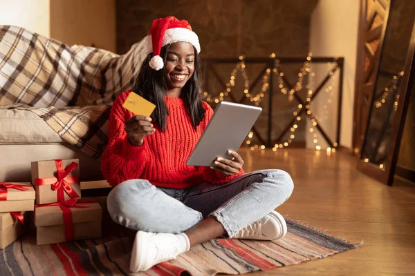 オンライン休日の販売。Santaの幸せな黒の女性帽子を使用してタブレットとクレジットカードでオンラインでクリスマスプレゼントを購入する家から — ストック写真