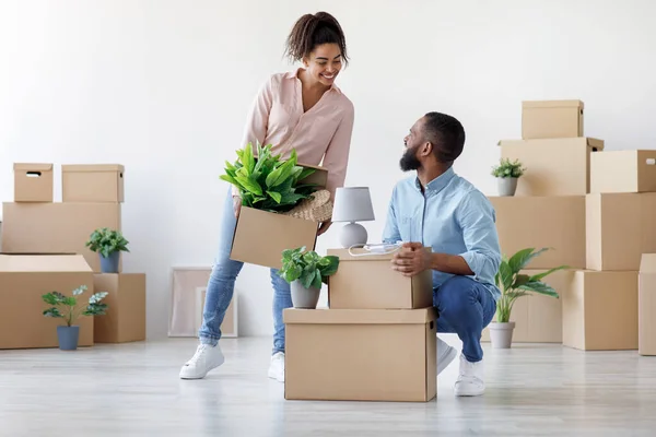 Sorrindo millennial marido preto e esposa descompactar caixas de papelão no quarto com paredes brancas e plantas envasadas — Fotografia de Stock