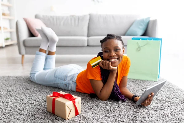 Jovencita alegre negro tumbado en el suelo con tableta y tarjeta de crédito, compras de regalos de vacaciones en línea desde casa —  Fotos de Stock