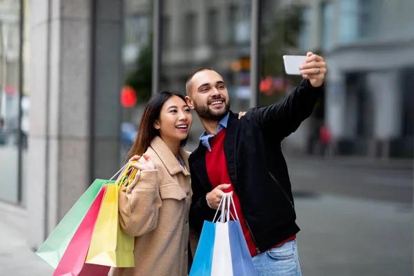 Wesoła para wielorasowa z torbami shopper robienie selfie na smartfonie przed dużym supermarketem — Zdjęcie stockowe