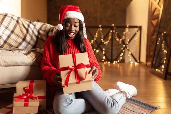 Festeggiamenti di Natale e Capodanno. Eccitato giovane donna nera in Santa cappello apertura regali di Natale sul pavimento a casa — Foto Stock