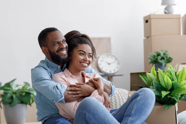 Satisfeito jovem negro marido e mulher abraçando, sentar no chão com caixas de papelão, coisas e plantas em novo apartamento — Fotografia de Stock