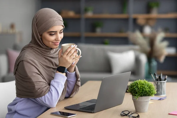 ผู้หญิงมุสลิมดูวิดีโอบนพีซีถือถ้วยดื่มกาแฟ — ภาพถ่ายสต็อก